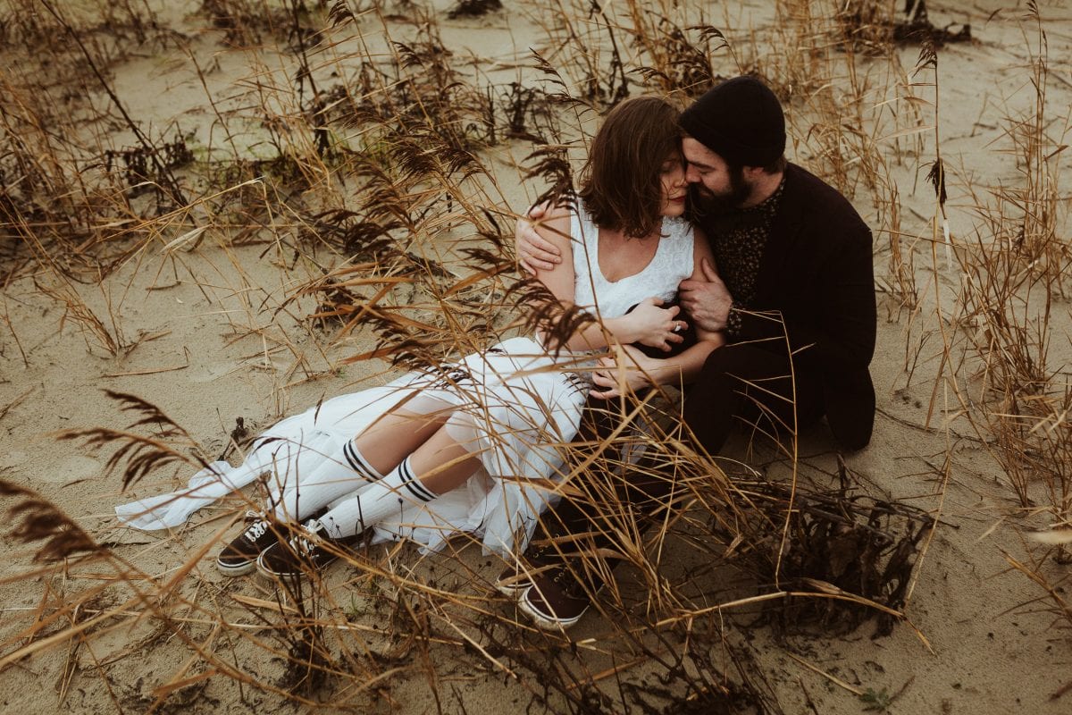 An eloping couple sitting in the sand, kissing. He's wearing a suit and a beanie, she's wearing a short designer's wedding dress, Vans sneakers and high skater's socks.