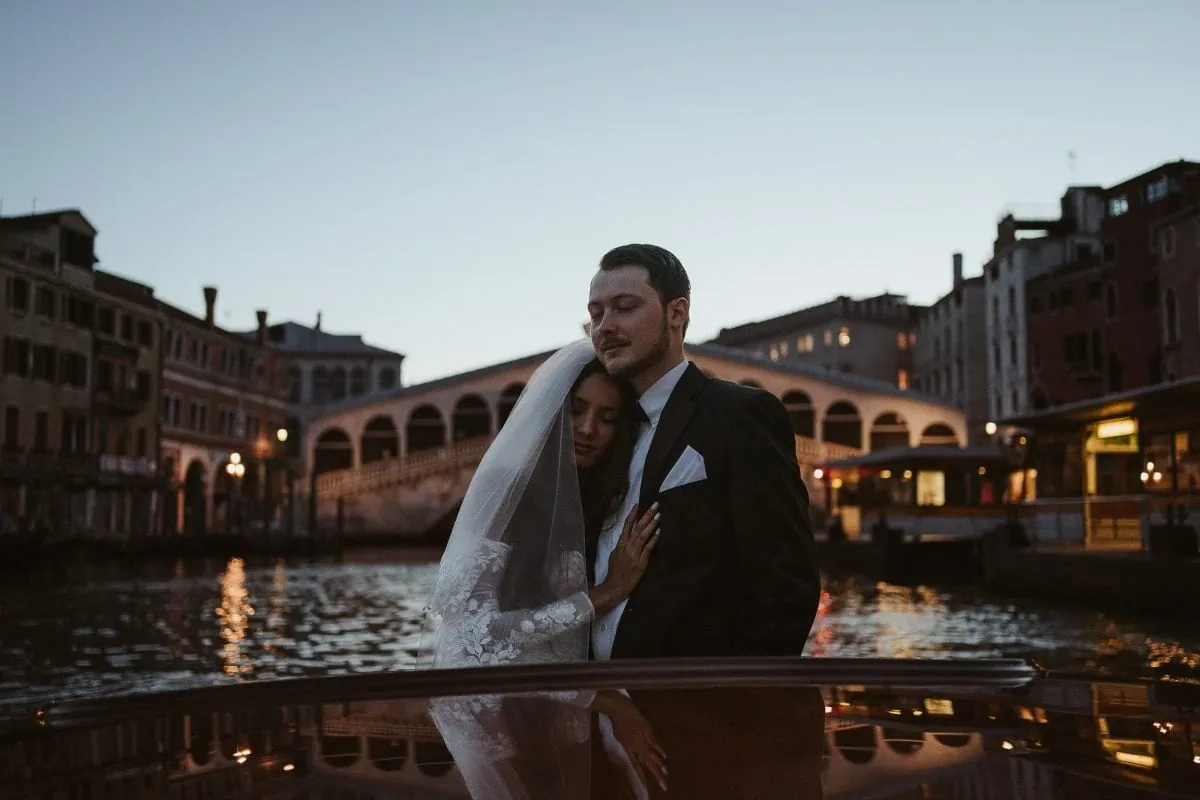 A luxuriously dressed couple on a motorboat in Venice, Italy during sunrise in front of the famous Rialto bridge on their elopement day.