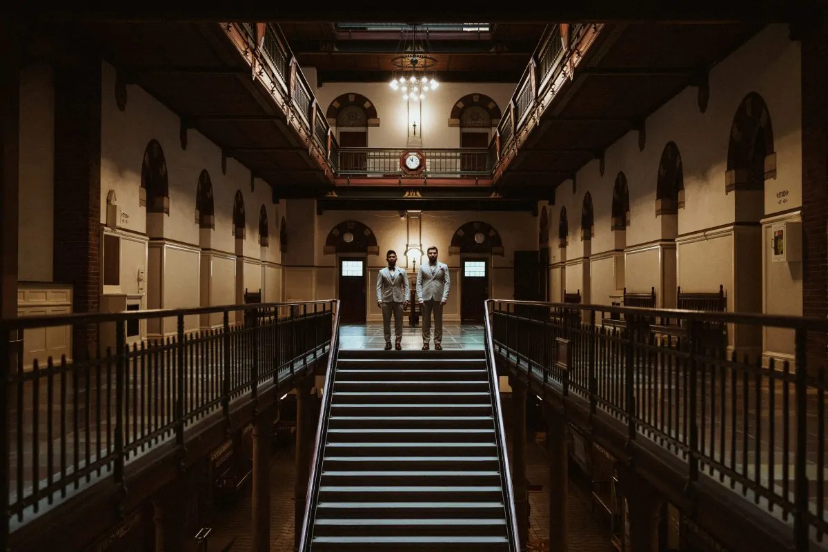 A gay couple in the town hall of Copenhagen right before their elopement ceremony. They're standing on top of a stairway. The scenery could be taken from a Wes Anderson movie.
