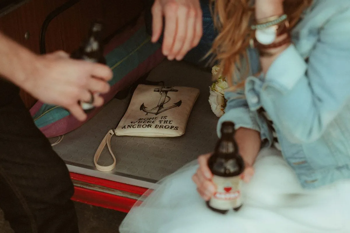 Close-up eines frisch verheirateten Paars, das sich beim Elopement in St. Peter-Ording ein Bier im Heck ihres Vans teilt. Neben ihnen liegt ein Canvas-Beutel mit der Aufschrift „Home is where the anchor drops“ – umgeben von sandigen Füßen, Jeansjacken und dieser besonderen Ruhe nach dem Ja-Wort.