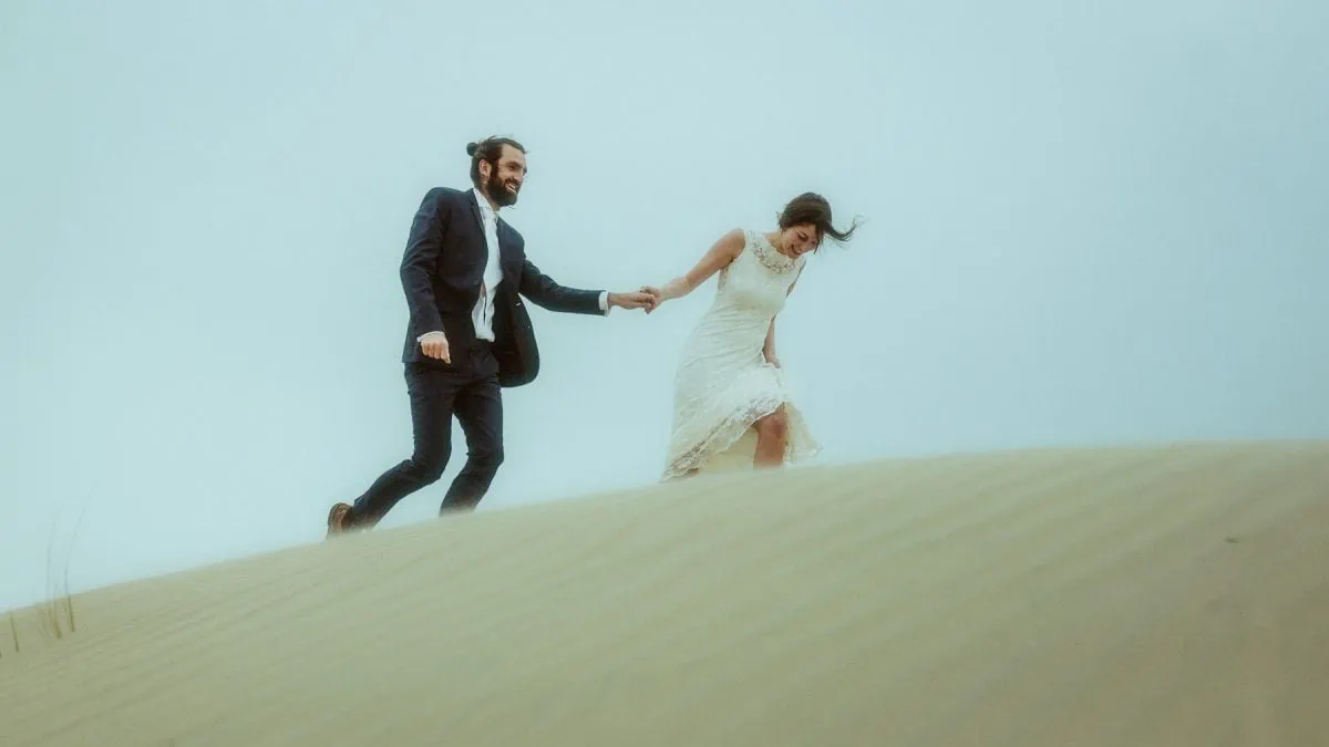 Ein Paar in Hochzeitskleidung rennt Hand in Hand über die windgepeitschten Sanddünen von Rømø, Dänemark – ihr Spitzenkleid fliegt im Wind, sein Jackett wird von der Brise erfasst. Ein ungezwungener Moment voller Lachen, Bewegung und wilder Nordsee-Energie.