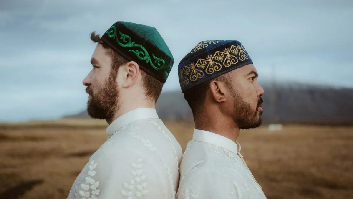 The image features a close side-by-side profile view of a gay couple wearing traditional embroidered tunics and ornate tubeteika hats, with a vast, open Icelandic landscape stretching out behind them.